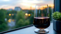 Glass of red wine on windowsill against blurred sunset cityscape with bokeh lights and greenery, creating romantic evening atmosphere for relaxation and celebration.
