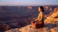 Young woman practicing meditation yoga pose on canyon cliff edge during golden hour sunrise with dramatic desert landscape background.