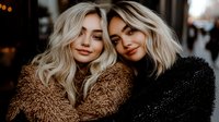 Two young Caucasian women with blonde wavy hair and natural makeup smiling at camera, wearing cozy textured winter coats in brown and black. Moody outdoor portrait.