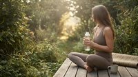 Woman with long brown hair, Caucasian ethnicity, sitting on wooden deck in lush forest holding water bottle, wearing athletic tank top, surrounded by green foliage and natural sunlight filtering through trees.