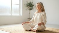 Peaceful middle-aged Caucasian woman meditating in lotus position wearing white clothing in bright minimalist room with plant for wellness and mindfulness.
