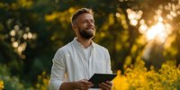 Smiling Caucasian man with beard holding tablet outdoors at sunset, surrounded by yellow wildflowers in natural park setting.
