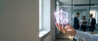 Businessman hand holding glowing shield icon with security symbol in modern office environment, representing data protection and cybersecurity solutions.
