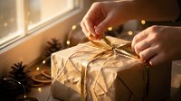 Hands wrapping golden twine around kraft paper gift box near window with warm string lights and holiday decorations for festive present preparation.