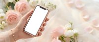 Female hand holding smartphone with blank screen surrounded by pink roses and white flowers on textured surface for app mockup and digital marketing.