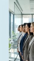 Diverse group of five professional women standing in modern office with glass partitions, looking toward windows with city view, wearing business casual attire for corporate teamwork concepts.