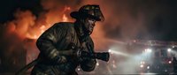 Firefighter in protective gear and helmet working at night emergency scene with dramatic smoke and fire truck lights in background.