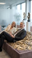 Confident senior Caucasian business couple sitting on pile of gold coins in modern office, symbolizing wealth, financial success, and retirement prosperity.