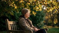 Senior Caucasian man in knit hat sitting peacefully on park bench during golden autumn afternoon, enjoying warm sunlight and fall foliage in tranquil outdoor setting.