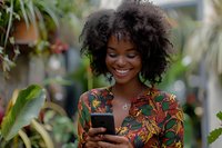 Young African American woman with natural curly hair smiling while using smartphone outdoors, wearing colorful floral pattern dress. Perfect for lifestyle, technology and positive emotion concepts.