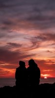 Silhouetted couple sitting together on rocky shore watching dramatic sunset with vibrant orange and red sky, perfect for romance, relationships, and travel marketing.