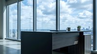Modern office reception desk with panoramic city skyline view through floor-to-ceiling windows, featuring minimalist black and gray design, potted plant, and coffee cup in contemporary high-rise workspace.