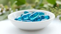 Blue gel capsules in white ceramic bowl on natural background with green foliage for health and wellness supplement marketing materials.