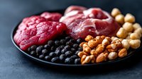 Fresh raw beef chunks with chickpeas, walnuts and black olives arranged on dark plate against black background, healthy protein-rich ingredients for cooking.