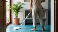 Woman in linen pants mopping turquoise tile floor in modern kitchen with palm plant and natural light streaming through wooden door frame.