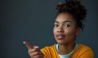 Young African American woman pointing finger at camera with confident expression wearing orange jacket against dark background for marketing.