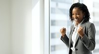 Young African American woman in professional business attire celebrating success with fist pump gesture near office window with city skyline view.