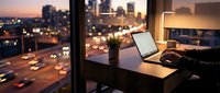 Modern office workspace with laptop computer on desk overlooking illuminated city skyline at sunset. Business professional remote work setup for productivity.