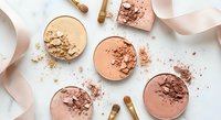 Crushed makeup powder compacts in nude and coral shades scattered with cosmetic brushes on white background for beauty and fashion concepts.
