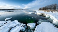 Dramatic winter landscape with floating ice chunks on dark water surface. Frozen lake or river with snow-covered shore and bare trees against cloudy blue sky.
