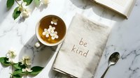 Flat lay of herbal tea with jasmine flowers, linen napkin with be kind text, open book and spoon on marble surface.