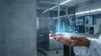 Young adult male with light skin tone wearing a white long-sleeved shirt, holding an open palm displaying a holographic data visualization with glowing blue charts and graphs in a modern office environment with glass partitions and fluorescent lighting.