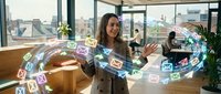 Young woman with long dark hair and medium skin tone, wearing a beige blazer, gestures toward holographic email and communication icons floating in air around her in a modern office with city skyline view, representing digital communication and business technology.