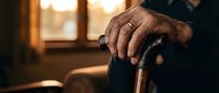 Elderly man's weathered hands gripping wooden walking cane in warm sunlit room, representing aging, independence, and senior care support at home.