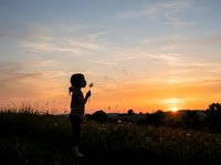 Young woman blowing dandelion seeds in meadow at golden sunset, silhouetted against warm sky with flying seeds and distant trees.