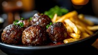 Delicious glazed meatballs served with golden french fries and fresh herbs on dark plate in restaurant setting for menu and food advertising.