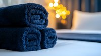 Rolled navy blue spa towels stacked on surface with soft bokeh lights in background creating relaxing wellness atmosphere for spa and hotel concepts.