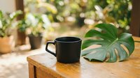 Black ceramic coffee mug on wooden table with monstera leaf and houseplants in bright natural light for morning routine and home decor concepts.