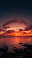 Dramatic sunset over calm ocean waters with vibrant orange and red sky, silhouetted clouds, and fishing boats on horizon. Rocky coastline in foreground. Ideal for travel, nature, and seascape projects.