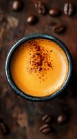 Creamy orange-hued drink topped with spice powder in dark ceramic cup, surrounded by scattered coffee beans on rustic surface, shot from above in moody lighting.