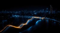 Aerial night cityscape with illuminated highway winding along waterfront, glowing blue skyscrapers reflecting in dark water, modern urban architecture creating dramatic scene.