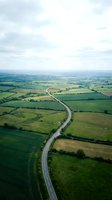 Aerial view of a straight highway cutting through patchwork farmland with green fields, hedgerows, and scattered trees under cloudy skies. Perfect for transportation, infrastructure, and rural landscape projects.