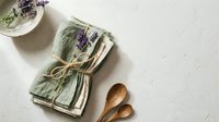 Rustic kitchen styling with sage green linen napkins, dried lavender sprigs, wooden spoons and ceramic bowl on textured white surface for cooking content.