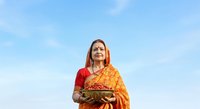 Woman of South Asian descent wearing traditional red and gold sari with dupatta, holding decorative plate, looking upward against blue sky background for cultural and festive content.