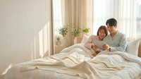 Happy Asian couple relaxing in bed using digital tablet together in bright modern bedroom with natural sunlight streaming through windows.