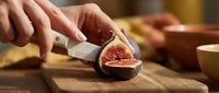 Young woman with fair complexion slicing fresh figs on wooden cutting board in warm kitchen setting. Hands preparing fruit for cooking or food styling purposes.