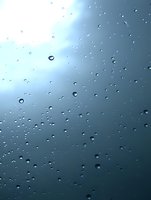 Raindrops on window glass against moody blue sky background create atmospheric mood with water droplets catching light and casting subtle shadows.