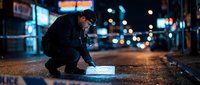 Young male detective examining evidence with flashlight on urban street at night during criminal investigation scene with city lights and traffic in background.