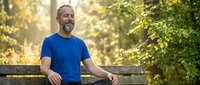 Middle-aged Caucasian man with beard wearing blue t-shirt sitting on wooden bench in sunny forest park setting, relaxing outdoors among green trees and natural light.