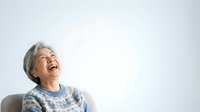 Senior woman with gray hair wearing cozy knit sweater, laughing joyfully while looking upward against white background. Suitable for healthcare, wellness, lifestyle and aging positively concepts.