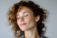 Young Caucasian woman with freckles and curly brown hair smiling with eyes closed in peaceful expression against light background, natural beauty portrait.