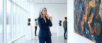 Professional businesswoman in navy suit talking on phone in modern art gallery with colorful abstract paintings and large windows creating bright atmosphere.