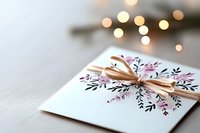 Elegant white greeting card with delicate floral pattern and golden bow against soft bokeh lights background, creating romantic holiday atmosphere.