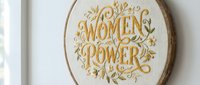 Women power embroidered on cream fabric in oval hoop with gold floral ornaments, isolated on white background for wall decor and feminist design projects.