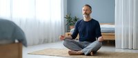 Middle-aged Caucasian man meditating in lotus position on beige carpet in bright modern living room. Wearing navy t-shirt and jeans. Peaceful mindfulness practice at home.