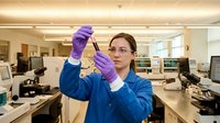 Young female scientist or laboratory technician of mixed ethnicity wearing blue lab coat and purple protective gloves, examining test tube in modern research facility with computers and equipment.